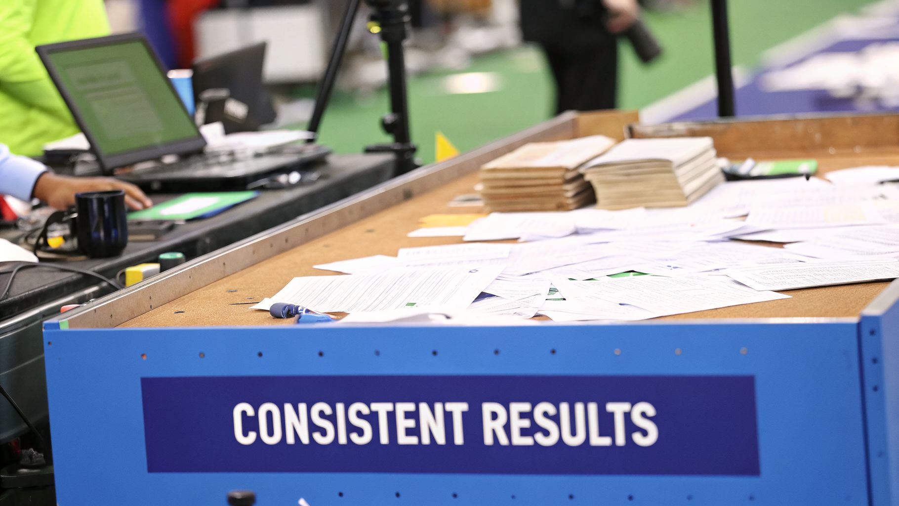 A busy desk with papers, a laptop, and a sign stating "CONSISTENT RESULTS."