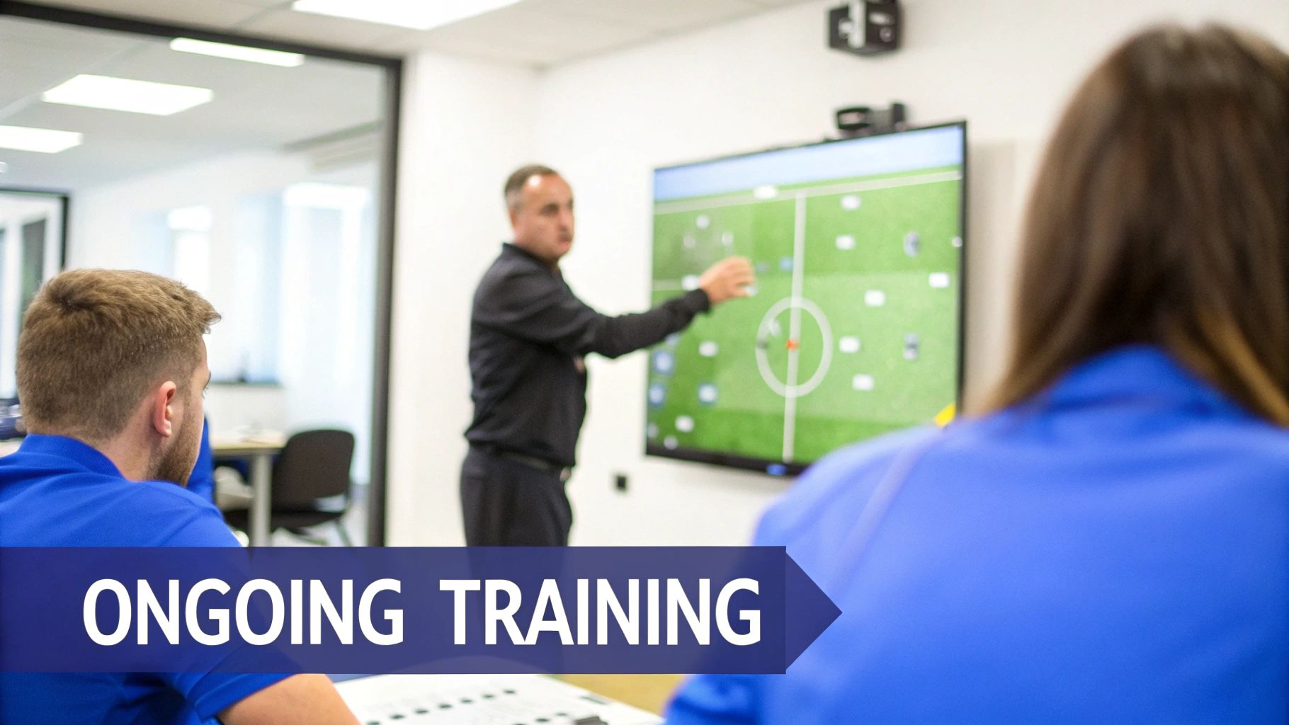An instructor points at a large screen displaying soccer tactics to students in a modern training room.