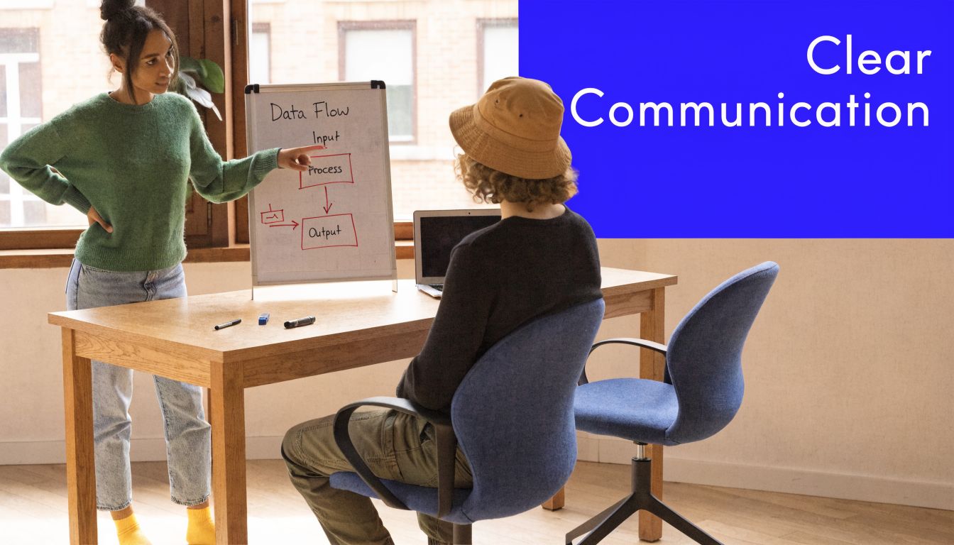 A young woman explains a data flow diagram on a whiteboard to a colleague sitting at a desk.
