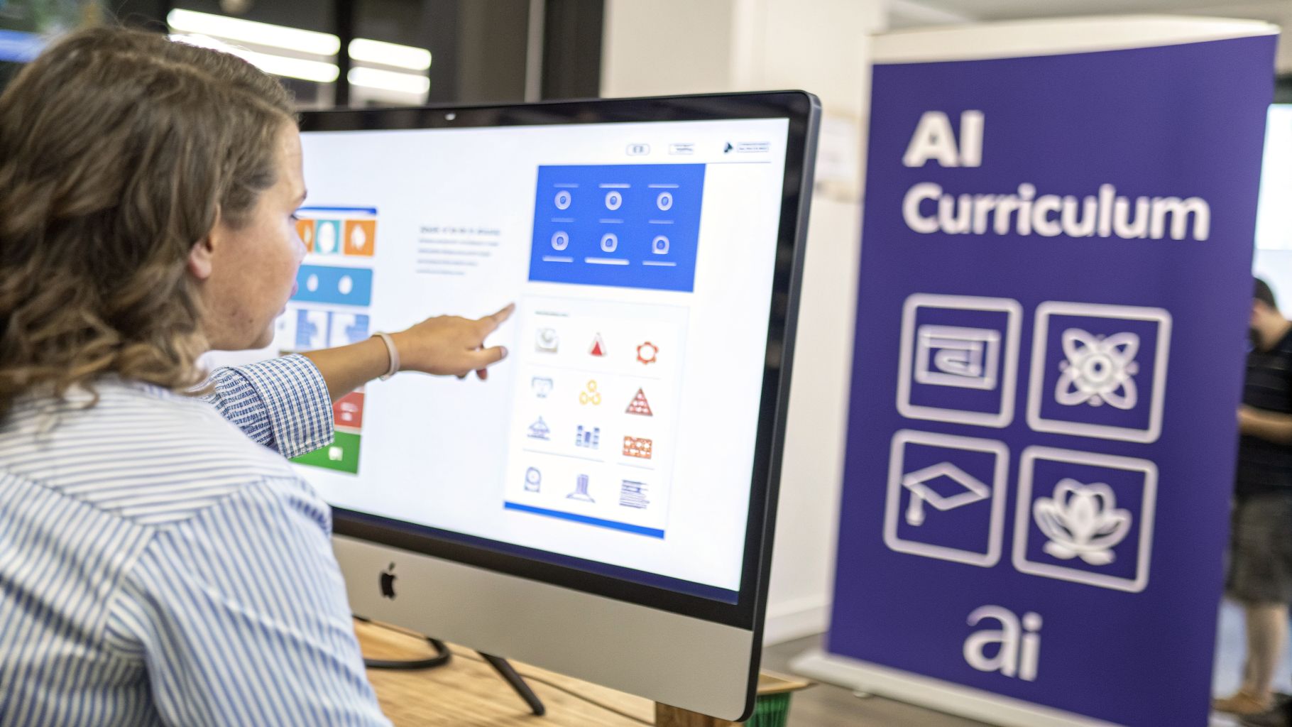 A person points at an iMac screen displaying an AI curriculum interface with various icons.