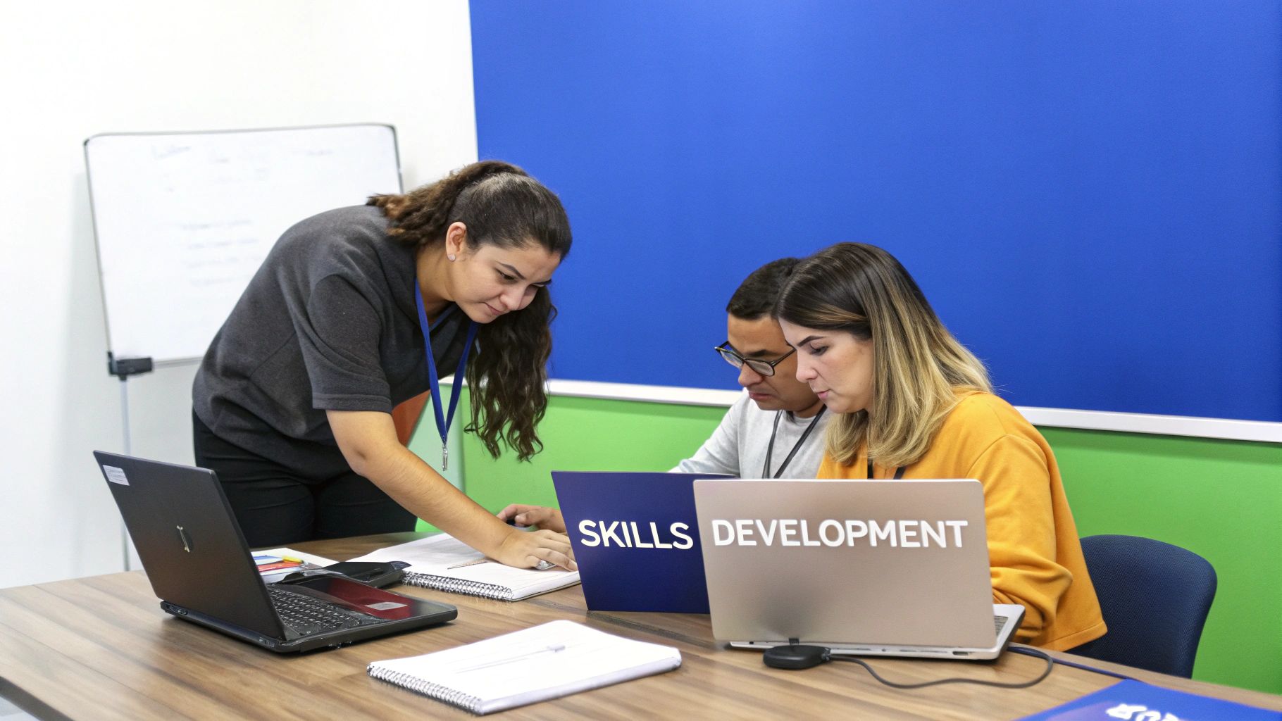 An instructor assists two students working on laptops displaying 'Skills Development' in a bright classroom.