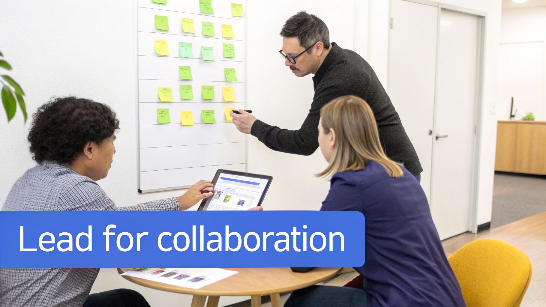 Three people collaborate in an office, one writing on a sticky note board, two discussing a tablet.