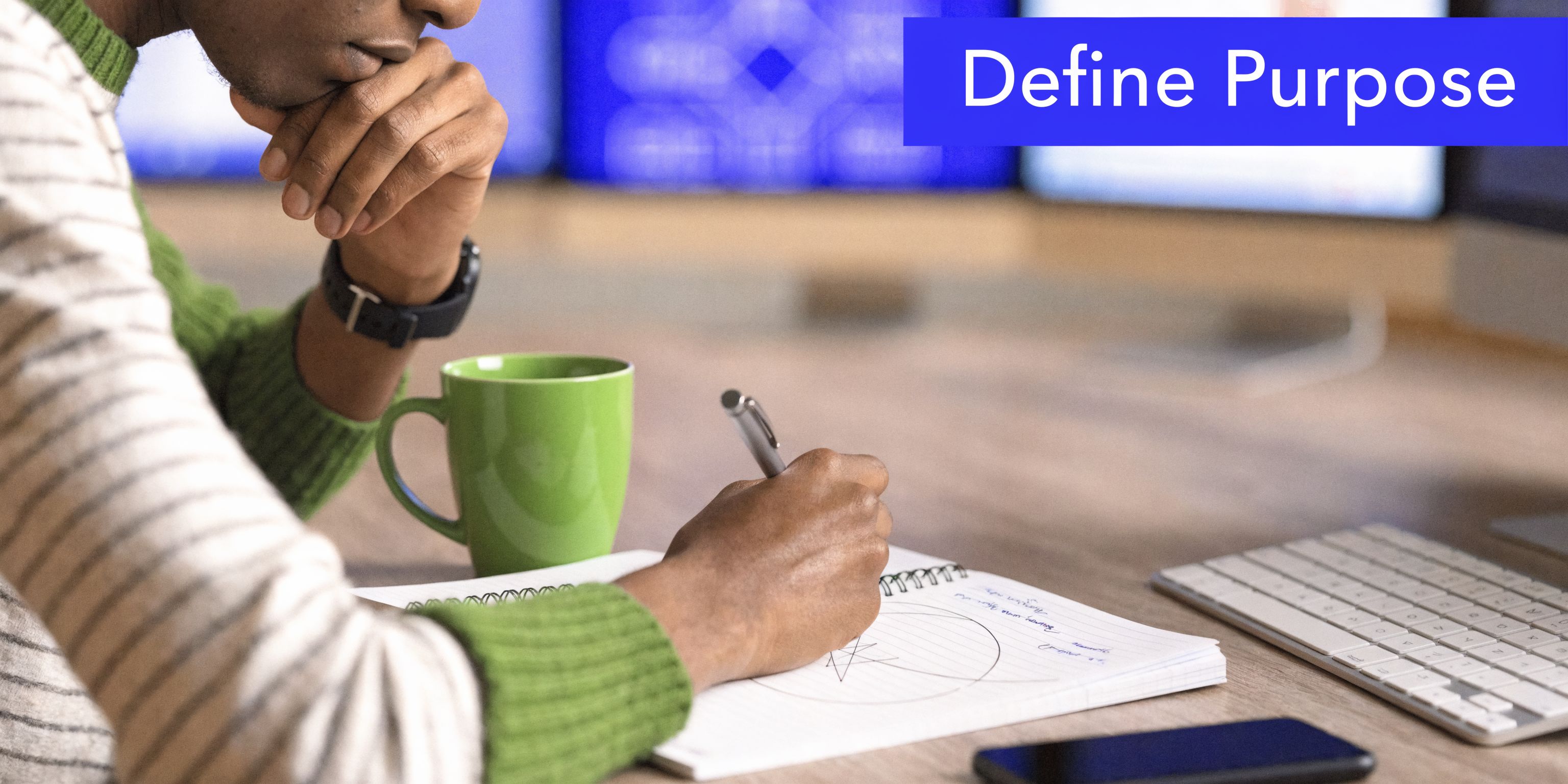 A person writing in a notebook at a desk with a coffee mug and computer screen.