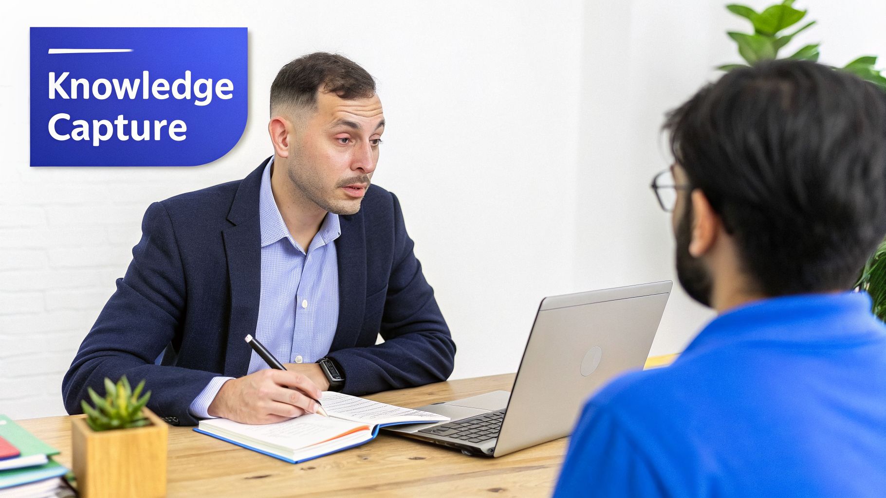 Two men collaborating at a desk, one taking notes while the other speaks, with a 'Knowledge Capture' sign.